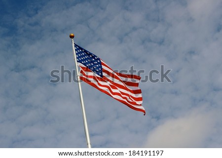 Patriotic waving red, white, and blue flag against a cloudy bright blue sky on a sunny afternoon.