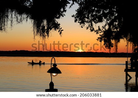 Fiery evening summer sunset at the lake with silhouetted canoe with fisherman in the background and a bright lit lamp and bulb in the foreground with tree branches touching down.