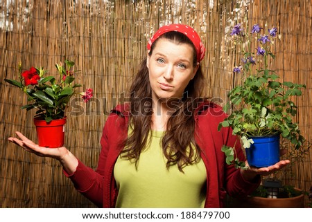 Woman gardening and holding two flower pots with different plants in front of a bamboo fence unable to choose one.