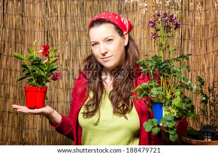 Woman in gardening holding two flower pots with different plants in front of a bamboo fence.