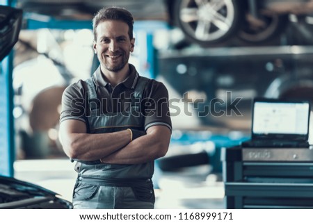 Handsome Smiling Car Machanic Standing in Garage. Portrait of Caucasian Adult Technician Master in Dark Uniform with Crossed Arms in Front of Automobile. Repairing Car Service Concept