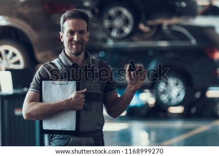 Auto Mechanic Holding Clipboard and Car Keys. Repair service. Handsome Serious Caucasian Man in Grey Uniform Standing in Front of Black Car and Finished Checkup of Service Order Working in Garage
