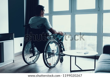 Disabled Person Sits in Wheelchair Against Window. Serious Sad Caucasian Man Wearing Casual Clothes and Look at Large Panoramic View in Bright Modern Living Room or Hospital.