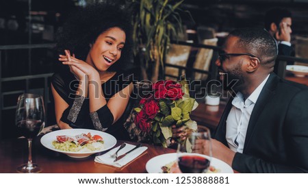 African American Couple Dating in Restaurant. Romantic Couple in Love Dating. Cutel Man and Girl in a Restaurant Making Order. Romantic Concept. Man Giving Bouquet of Flowers. Red Roses.