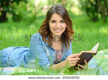 Beautiful young woman-student reading book on grass, against background of summer green park.