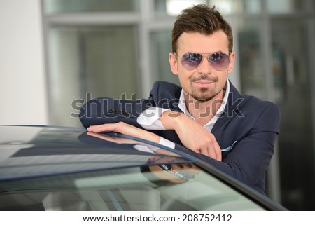 Confident man in formalwear leaning at the car and looking at camera while standing at car dealership
