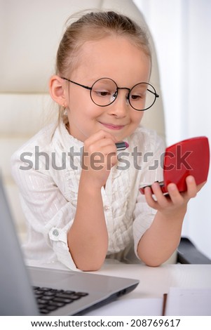 Little business woman. Cute girl with glasses and a solemn sitting at the table, paints lips with lipstick