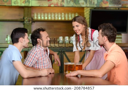 Beer for gentlemen. Beautiful young waitress serving beer while three men sitting at the table in beer pub
