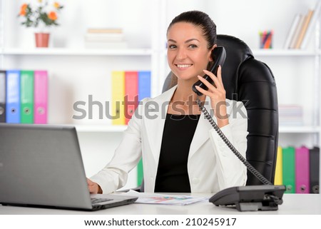 Talking at phone. Confident middle-aged businesswoman sitting at her working place and looking over shoulder