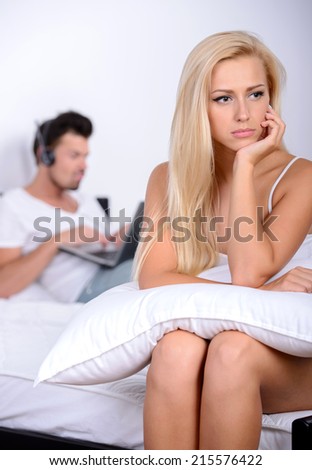 Problems in the family. Angry young woman sitting on the bed, against her husband, who plays the game on the computer