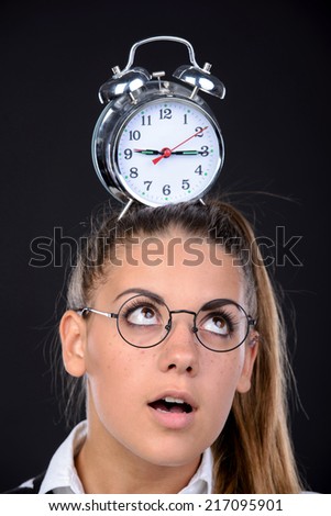 Young nerd woman crazy expression in glasses, holding an alarm clock on head on black background
