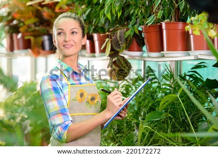 Working in greenhouse. Beautiful woman in uniform writing something in her note pad and looking at the flowers while standing in greenhouse