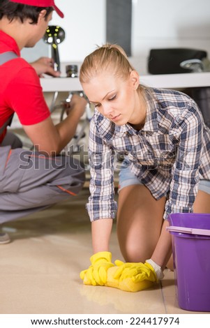 Young woman is cleaning the floor in the bathroom, while plumber repairs the pipes.
