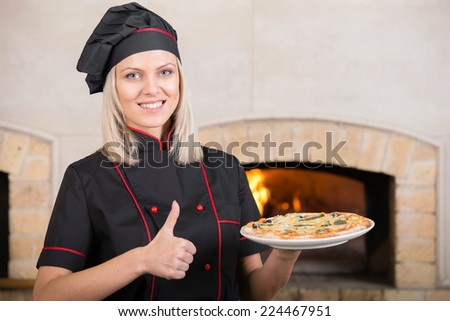 Woman chef is standing at the kitchen entrance with the wood oven in the background with pizza.