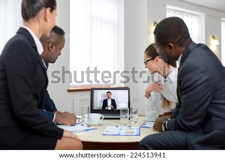 Group of male and female businesspeople seated at a table watching an online conference on a computer screen