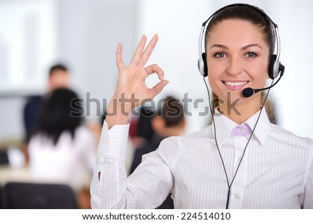 Female customer support operator with headset and smiling, people group in background at modern bright office indoors
