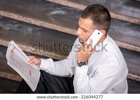 Young unemployed man is looking for a job in the newspaper,  and phone, sitting on the stairs.