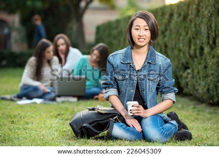 Portrait of a young, asian, smiling female student, with blurred students are sitting in the park. She is looking at the camera.