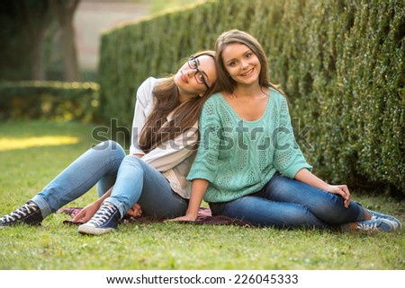 Two smiling female students are sitting on the grass at campus. They are looking at camera.