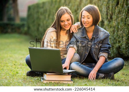 Two smiling female students are sitting on the grass. They are working on laptop at campus.