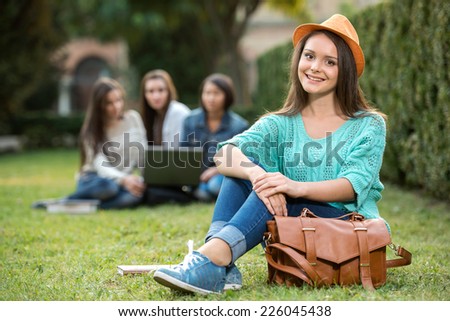 Portrait of a young, smiling female student, with blurred students are sitting in the park. She is looking at the camera.