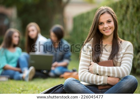 Portrait of a smiling college girl is holding book with blurred students are sitting in the park.