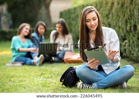 Portrait of a smiling college girl is holding tablet PC with blurred students are sitting in the park.