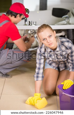 Woman is cleaning the floor in the bathroom, while plumber repairs the pipes.
