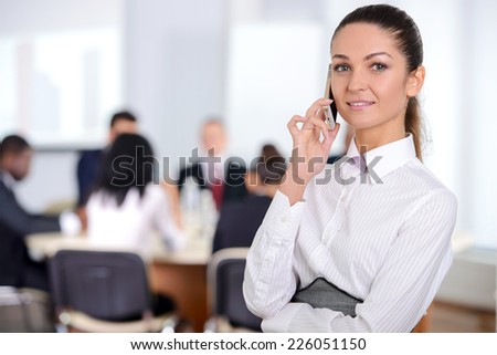 Business woman, talking on the phone, with her staff, people group in background at modern bright office indoors