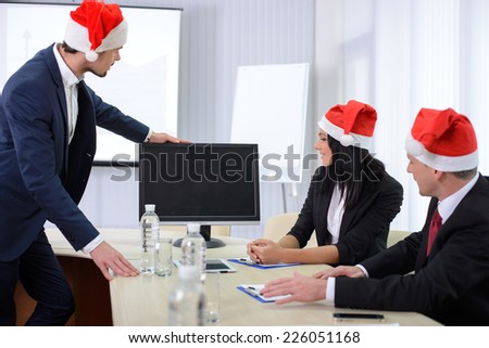 Group of male and female businesspeople seated at a table watching an online conference on a computer screen, during the celebration of Christmas