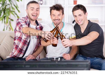 Three young football fans are cheering football match at home and drinking beer.