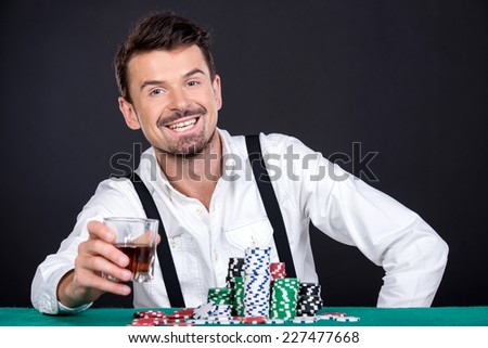 Happy man is playing poker and drinking whiskey in casino. Gambling chips on the green table.