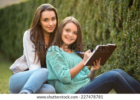 Two smiling female students are sitting on the grass with book, at campus. They are looking at camera.