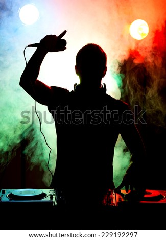 Silhouette of a young DJ at work with a microphone and the club lights on the background.