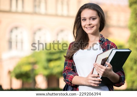 Portrait of a young beautiful student with university building in the background. She is holding books in hands.