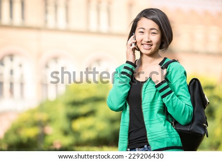 Portrait of a young smiling asian student with university building in the background. She is saying by phone.