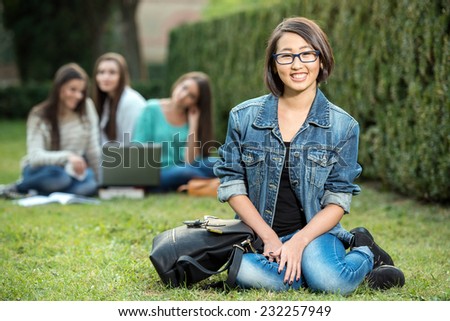 Portrait of a young, asian, smiling female student, with blurred students are sitting in the park. She is looking at the camera.
