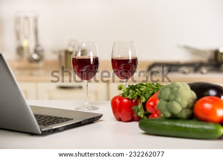 Two glasses of wine vegetables and computer on a table,  in the kitchen.