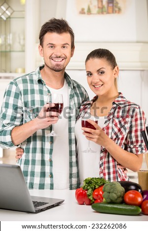 Young beautiful couple are drinking wine in the kitchen, vegetables and computer on a table.