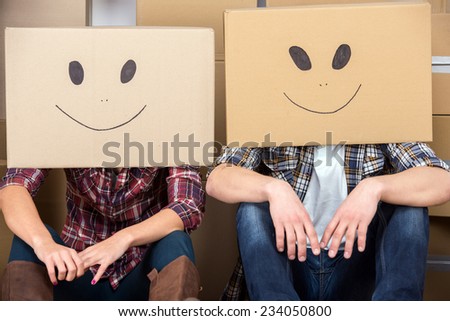 Couple with cardboard boxes on their heads with smiley face are sitting on floor after the moving house.