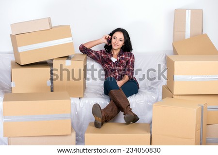 Woman in a new home with cardboard boxes, is sitting on covered sofa and speaking by phone.