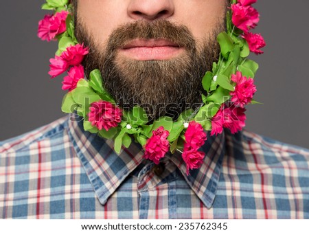 Close-up of man with flowers in his beard is standing against grey background.