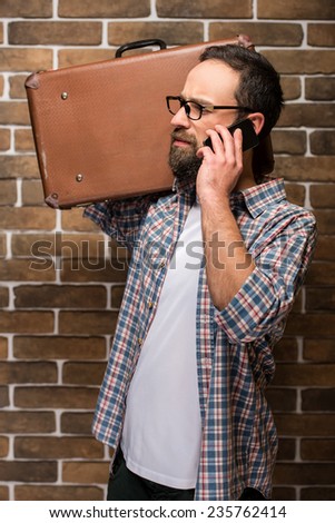 Young bearded man with a suitcase on his shoulder and speaking by phone while standing against brick wall.