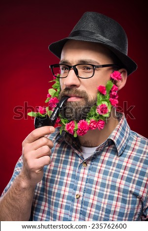 Brutal man with flowers in his beard, in hat and with pipe is standing against red background.