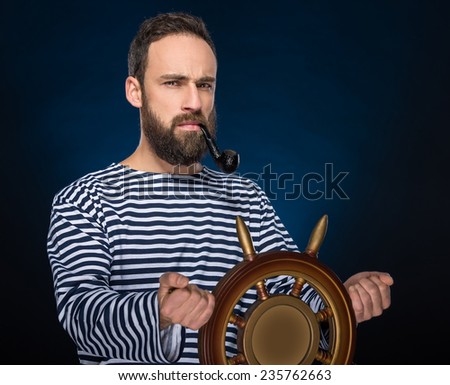 Young bearded sailor in striped clothing is smoking a pipe while standing with steering wheel.