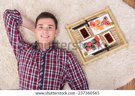 Young man with sushi is lying on the floor at home. He is looking at the camera. Top view.