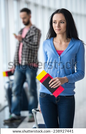 Portrait of young woman with passport and luggage in airport. Young man on background.
