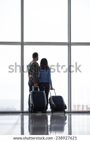 Young couple with suitcases are looking through airport window while waiting for flight. Back view.