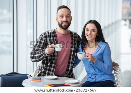 Young couple are  sitting with suitcase in international airport, are drinking coffee and looking at the camera.