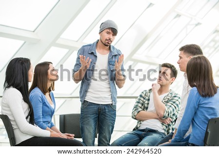 Young man is sharing his problems with people. View of man is telling something and gesturing while group of people are sitting in front of him and listening.